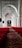 An interior view of a mosque corridor featuring elegant architectural arches. Ornate chandeliers hang from the ceiling, casting a warm glow on the surroundings. The floor is covered with a richly patterned red carpet extending into the distance.