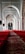 An interior view of a mosque corridor featuring elegant architectural arches. Ornate chandeliers hang from the ceiling, casting a warm glow on the surroundings. The floor is covered with a richly patterned red carpet extending into the distance.