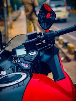A close-up of a motorcycle handlebar showcasing a vibrant red and black design. The focus is on the rearview mirror and key chain with a blurred street scene in the background.