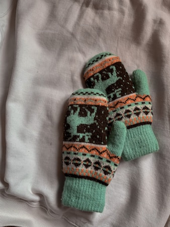 A pair of knitted mittens adorned with reindeer and geometric patterns lies on a soft, white fabric background. The mittens feature a mix of turquoise, brown, orange, and white colors with intricate designs.