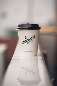 A paper coffee cup with a black lid sits on a reflective surface, displaying the logo 'Upsolute Coffee'. The background is softly blurred, giving a sense of depth and a focus on the cup. The cup's reflection is visible on the surface beneath.