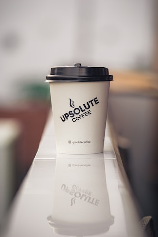 A paper coffee cup with a black lid sits on a reflective surface, displaying the logo 'Upsolute Coffee'. The background is softly blurred, giving a sense of depth and a focus on the cup. The cup's reflection is visible on the surface beneath.