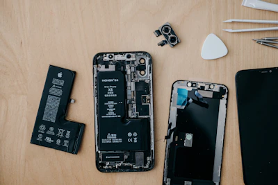 Various components of a disassembled smartphone are spread out on a wooden surface. Visible parts include the phone battery, camera module, front screen panel, and several small repair tools. The disassembled phone reveals intricate internal electronics.