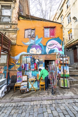 A vibrant street scene featuring a small art shop with an orange facade. The exterior wall is adorned with colorful murals, including large eyes and abstract faces. The shop displays an array of artworks, including paintings and framed pieces. A person in a green jacket is arranging items outside the shop. The cobblestone street and old buildings add to the rustic charm of the setting.