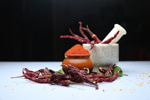 An arrangement featuring a stone mortar and pestle with dried red chili peppers both inside and surrounding it. A small brown pot filled with vibrant red chili powder sits in front, while loose chili seeds are scattered around on a light blue surface.