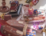 An arrangement of various decorative and craft items on a surface, including a toy model truck, glass tubes filled with colored sequins, a small glass jar with a cork lid containing pink sequins, small wrapped candies, wooden blocks with letters, and a gift box with a satin ribbon. The scene is colorful and has a playful, whimsical feel.