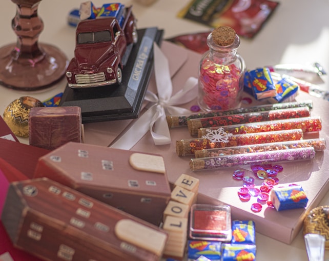An arrangement of various decorative and craft items on a surface, including a toy model truck, glass tubes filled with colored sequins, a small glass jar with a cork lid containing pink sequins, small wrapped candies, wooden blocks with letters, and a gift box with a satin ribbon. The scene is colorful and has a playful, whimsical feel.