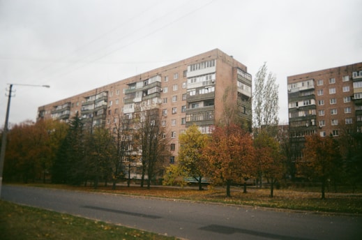 A row of mid-rise apartment buildings made of brick and concrete, with multiple balconies. In the foreground, there are several trees with colorful autumn foliage in shades of yellow, orange, and red. The road in front of the buildings appears empty, and the sky is overcast, creating a muted atmosphere.