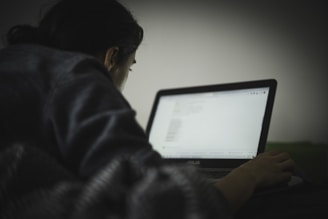 A person is lying down on a bed or couch using a laptop. The lighting is dim, with most of the environment shaded, giving a sense of privacy or late-night work. The focus is on the screen of the laptop, which appears to have text on it, suggesting the person is reading or coding.