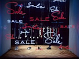 A shop window display featuring multiple neon signs with the word 'Sale' in red and blue. Inside the window, several items of clothing on hangers are visible, each tagged with a sale price. The floor has a herringbone wooden pattern and a few pairs of shoes are placed on it.