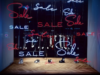 A shop window display featuring multiple neon signs with the word 'Sale' in red and blue. Inside the window, several items of clothing on hangers are visible, each tagged with a sale price. The floor has a herringbone wooden pattern and a few pairs of shoes are placed on it.