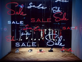 A shop window display featuring multiple neon signs with the word 'Sale' in red and blue. Inside the window, several items of clothing on hangers are visible, each tagged with a sale price. The floor has a herringbone wooden pattern and a few pairs of shoes are placed on it.