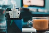 A box of cigarettes with a bright blue and green design is open, displaying several cigarettes with white and blue filters. In the background, a glass cup filled with a light brown beverage is placed on a table. The setting appears to be indoors, with a blurry scene on a monitor or television and soft lighting creating bokeh effects.