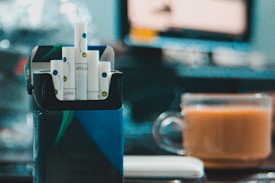 A box of cigarettes with a bright blue and green design is open, displaying several cigarettes with white and blue filters. In the background, a glass cup filled with a light brown beverage is placed on a table. The setting appears to be indoors, with a blurry scene on a monitor or television and soft lighting creating bokeh effects.