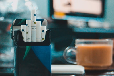 A box of cigarettes with a bright blue and green design is open, displaying several cigarettes with white and blue filters. In the background, a glass cup filled with a light brown beverage is placed on a table. The setting appears to be indoors, with a blurry scene on a monitor or television and soft lighting creating bokeh effects.