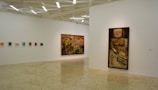 A minimalistic art gallery with white walls displays several pieces of modern art. Smaller framed artworks are aligned on the left wall, while two large paintings are prominently showcased on the right and central walls. The flooring is neutral-toned, and the ceiling features geometric light fixtures providing even illumination.