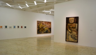 A minimalistic art gallery with white walls displays several pieces of modern art. Smaller framed artworks are aligned on the left wall, while two large paintings are prominently showcased on the right and central walls. The flooring is neutral-toned, and the ceiling features geometric light fixtures providing even illumination.