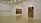 A minimalistic art gallery with white walls displays several pieces of modern art. Smaller framed artworks are aligned on the left wall, while two large paintings are prominently showcased on the right and central walls. The flooring is neutral-toned, and the ceiling features geometric light fixtures providing even illumination.