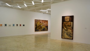 A minimalistic art gallery with white walls displays several pieces of modern art. Smaller framed artworks are aligned on the left wall, while two large paintings are prominently showcased on the right and central walls. The flooring is neutral-toned, and the ceiling features geometric light fixtures providing even illumination.