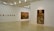 A minimalistic art gallery with white walls displays several pieces of modern art. Smaller framed artworks are aligned on the left wall, while two large paintings are prominently showcased on the right and central walls. The flooring is neutral-toned, and the ceiling features geometric light fixtures providing even illumination.