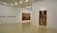 A minimalistic art gallery with white walls displays several pieces of modern art. Smaller framed artworks are aligned on the left wall, while two large paintings are prominently showcased on the right and central walls. The flooring is neutral-toned, and the ceiling features geometric light fixtures providing even illumination.
