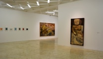 A minimalistic art gallery with white walls displays several pieces of modern art. Smaller framed artworks are aligned on the left wall, while two large paintings are prominently showcased on the right and central walls. The flooring is neutral-toned, and the ceiling features geometric light fixtures providing even illumination.