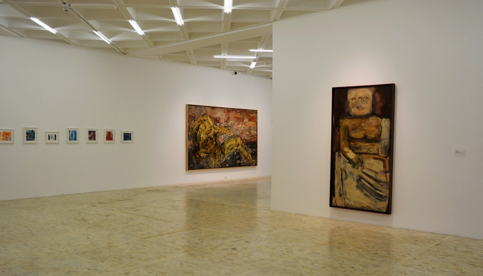 A minimalistic art gallery with white walls displays several pieces of modern art. Smaller framed artworks are aligned on the left wall, while two large paintings are prominently showcased on the right and central walls. The flooring is neutral-toned, and the ceiling features geometric light fixtures providing even illumination.