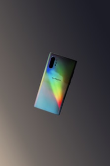 A Samsung smartphone with a glossy back surface reflecting a spectrum of rainbow colors is centered against a gradient dark background. The phone's design is sleek and modern, with multiple camera lenses visible at the top left.