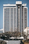 A tall, multi-story hotel building with a modern architectural design stands prominently. The structure is symmetrical with horizontal lines, large windows, and a central vertical column. Trees and a person can be seen in the foreground, adding a touch of greenery to the urban setting.
