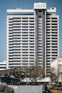 A tall, multi-story hotel building with a modern architectural design stands prominently. The structure is symmetrical with horizontal lines, large windows, and a central vertical column. Trees and a person can be seen in the foreground, adding a touch of greenery to the urban setting.