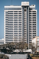 A tall, multi-story hotel building with a modern architectural design stands prominently. The structure is symmetrical with horizontal lines, large windows, and a central vertical column. Trees and a person can be seen in the foreground, adding a touch of greenery to the urban setting.