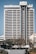 A tall, multi-story hotel building with a modern architectural design stands prominently. The structure is symmetrical with horizontal lines, large windows, and a central vertical column. Trees and a person can be seen in the foreground, adding a touch of greenery to the urban setting.