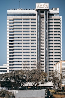 A tall, multi-story hotel building with a modern architectural design stands prominently. The structure is symmetrical with horizontal lines, large windows, and a central vertical column. Trees and a person can be seen in the foreground, adding a touch of greenery to the urban setting.