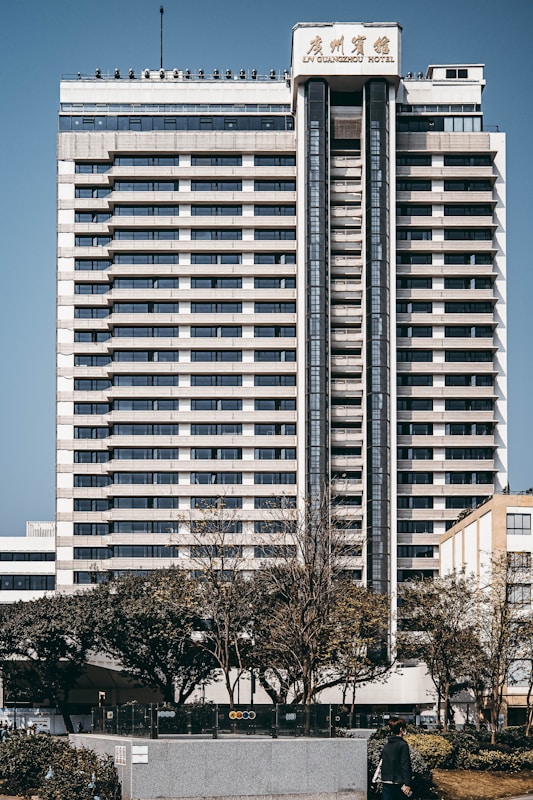 A tall, multi-story hotel building with a modern architectural design stands prominently. The structure is symmetrical with horizontal lines, large windows, and a central vertical column. Trees and a person can be seen in the foreground, adding a touch of greenery to the urban setting.
