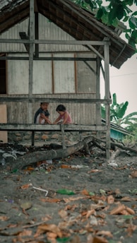 Two children are sitting on a wooden platform in front of a rustic building, surrounded by sand and scattered leaves. A large tree branch is visible nearby, and the building is made of wooden planks with a slanted roof. In the background, lush green foliage can be seen.