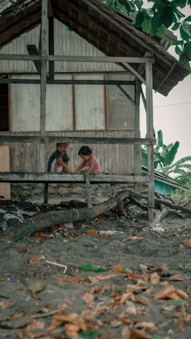 Two children are sitting on a wooden platform in front of a rustic building, surrounded by sand and scattered leaves. A large tree branch is visible nearby, and the building is made of wooden planks with a slanted roof. In the background, lush green foliage can be seen.