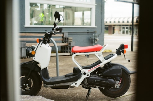 A small, modern scooter with a white frame and a bright red seat is parked in front of a grey building. The scooter has a compact design with black wheels and orange reflectors on the front. In the background, there is a large window and some concrete blocks, suggesting an urban environment.