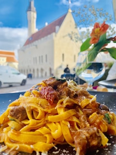 A plate of pasta with meat, garnished with pieces of tomato and cheese, sits prominently in the foreground. In the background, a historic building with a red roof and a tall spire rises against a bright blue sky. A glass of water and a vase with an orange flower are visible, adding to the composition.