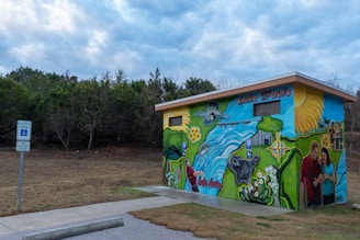 A small building with vibrant murals depicting nature scenes, animals, people enjoying outdoor activities, and the names 'Lake Travis' and 'Lake Austin'. The structure is set against a backdrop of trees under a cloudy sky, with a sign indicating reserved parking for accessibility nearby.