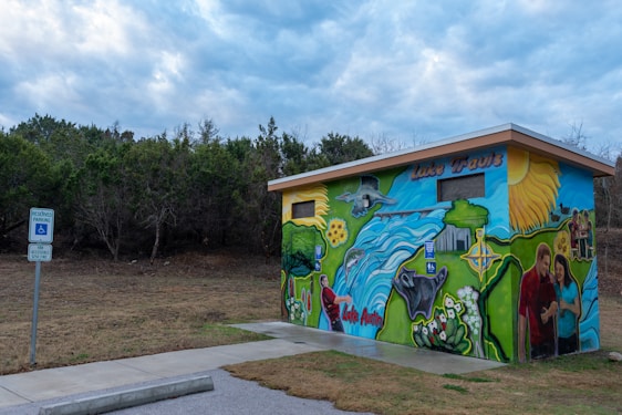 A small building with vibrant murals depicting nature scenes, animals, people enjoying outdoor activities, and the names 'Lake Travis' and 'Lake Austin'. The structure is set against a backdrop of trees under a cloudy sky, with a sign indicating reserved parking for accessibility nearby.