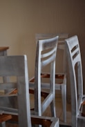 A group of unassembled or partially assembled wooden chairs is arranged in a room with a plain beige wall. The chairs are painted in a light, muted color that appears weathered or unfinished. The legs and backrests are visible, and there are angular wooden components featuring an orange-brown hue.