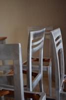 A group of unassembled or partially assembled wooden chairs is arranged in a room with a plain beige wall. The chairs are painted in a light, muted color that appears weathered or unfinished. The legs and backrests are visible, and there are angular wooden components featuring an orange-brown hue.