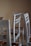 A group of unassembled or partially assembled wooden chairs is arranged in a room with a plain beige wall. The chairs are painted in a light, muted color that appears weathered or unfinished. The legs and backrests are visible, and there are angular wooden components featuring an orange-brown hue.