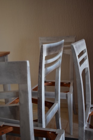 A group of unassembled or partially assembled wooden chairs is arranged in a room with a plain beige wall. The chairs are painted in a light, muted color that appears weathered or unfinished. The legs and backrests are visible, and there are angular wooden components featuring an orange-brown hue.
