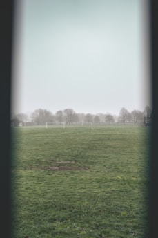 A foggy football field with a large expanse of green grass. In the background, there are several bare trees and two white soccer goals. The scene appears to be devoid of people, and the weather is overcast, contributing to a serene and slightly melancholic atmosphere.