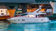 A luxury white yacht is sailing under a large concrete bridge in a bustling urban environment. Red buoys are attached to the sides of the yacht, and a person in blue clothing is visible on deck. The water is a vibrant blue, contrasting with the grey tones of the bridge structure above.