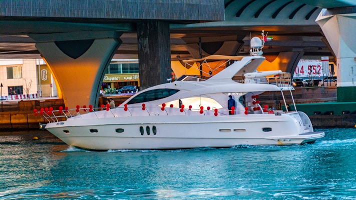 A luxury white yacht is sailing under a large concrete bridge in a bustling urban environment. Red buoys are attached to the sides of the yacht, and a person in blue clothing is visible on deck. The water is a vibrant blue, contrasting with the grey tones of the bridge structure above.