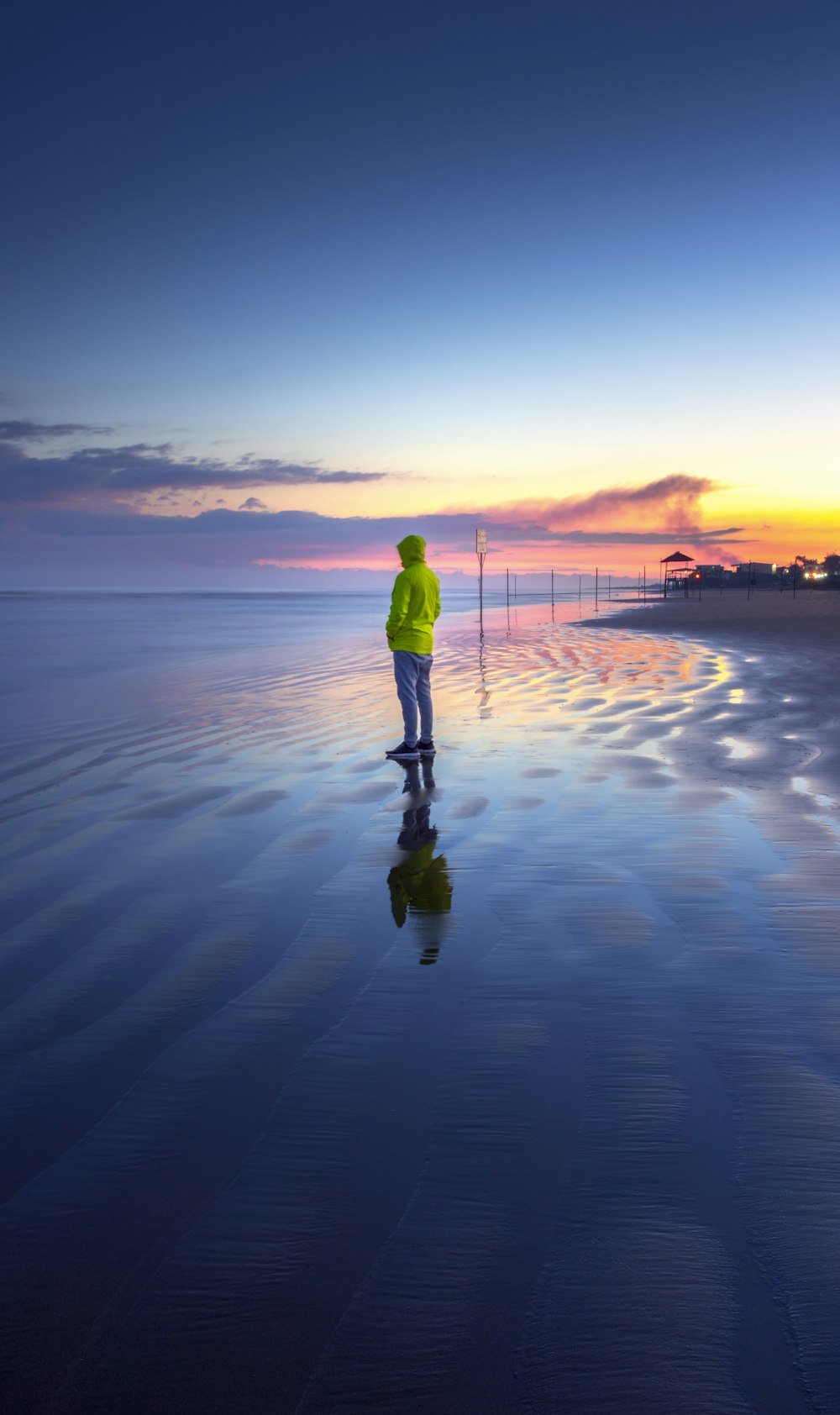 man in green jacket and black pants standing on seashore during sunset