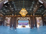 A traditional architectural interior features ornate wooden columns and beams adorned with intricate carvings. A large, decorative lantern with gold accents hangs from the ceiling. The floor is paved with polished stone tiles. In the background, there is a distant structure with purple and white decorations.