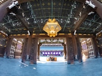 A traditional architectural interior features ornate wooden columns and beams adorned with intricate carvings. A large, decorative lantern with gold accents hangs from the ceiling. The floor is paved with polished stone tiles. In the background, there is a distant structure with purple and white decorations.